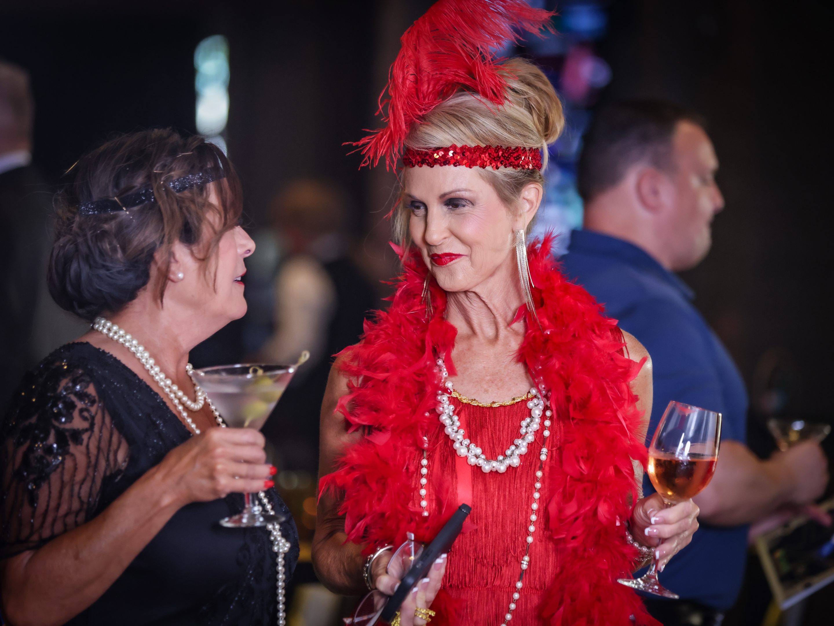 At the party, two women dressed in red and feathers are discussing, possibly affiliated with The LIME Foundation of Santa Rosa.