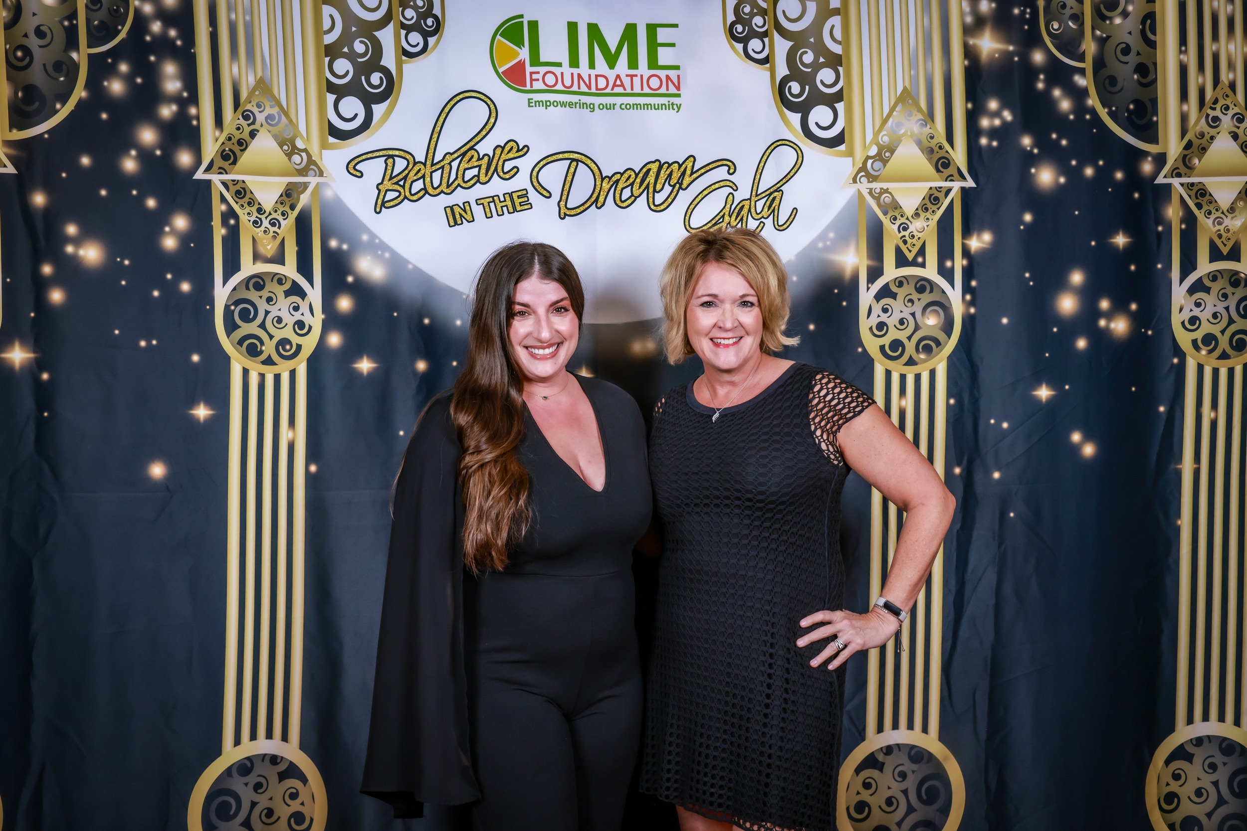 Two women posing for a photo in front of a golden backdrop at The LIME Foundation event.